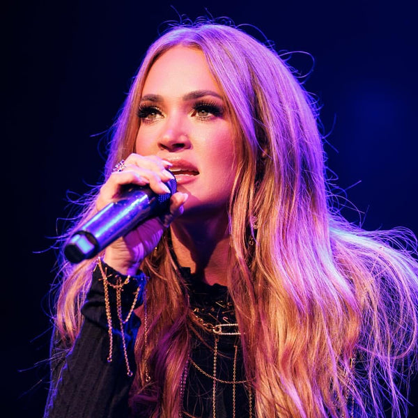 Person with long blonde hair singing into a microphone on a dark background