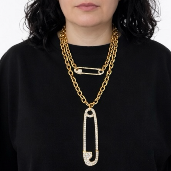 Person wearing a gold chain necklace with a prominent safety pin pendant on a black background