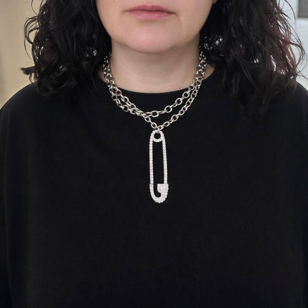 Person wearing a silver chain necklace with a prominent safety pin pendant on a black background