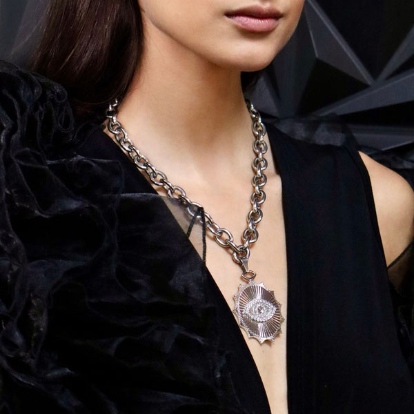 white gold plated large evil eye pendant hanging on a chunky chain black deep v neck dress brunette model