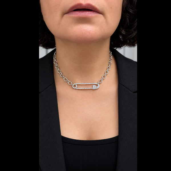 A close-up of a model wearing a silver safety-pin style chain necklace with a pavé-set heart-shaped center, styled with a black blazer.