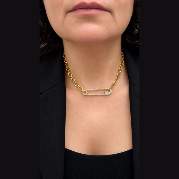 A close-up of a model wearing a gold chain necklace with a safety-pin-shaped pendant paved with clear stones, styled with a black blazer.