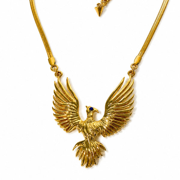 A gold-toned eagle pendant necklace with wings spread wide, featuring a small dark blue gemstone accent in the eye, shown on a white background.