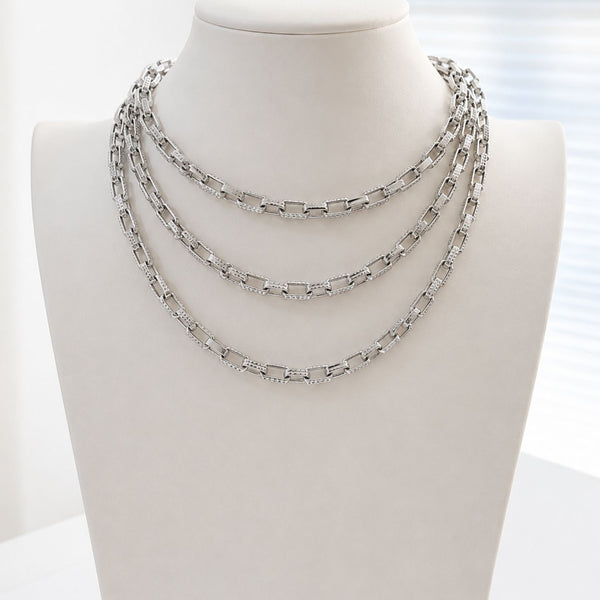 A three-layered silver chain necklace displayed on a white mannequin bust against a bright, softly lit background.