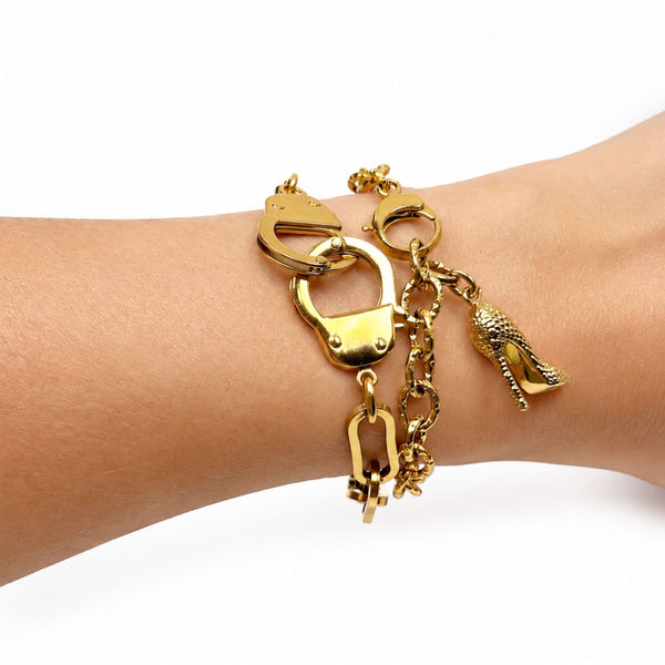 A gold-toned charm bracelet worn on a person's wrist, featuring a lobster clasp, chain links, a padlock charm, and a high-heeled shoe charm, shown against a plain white background.