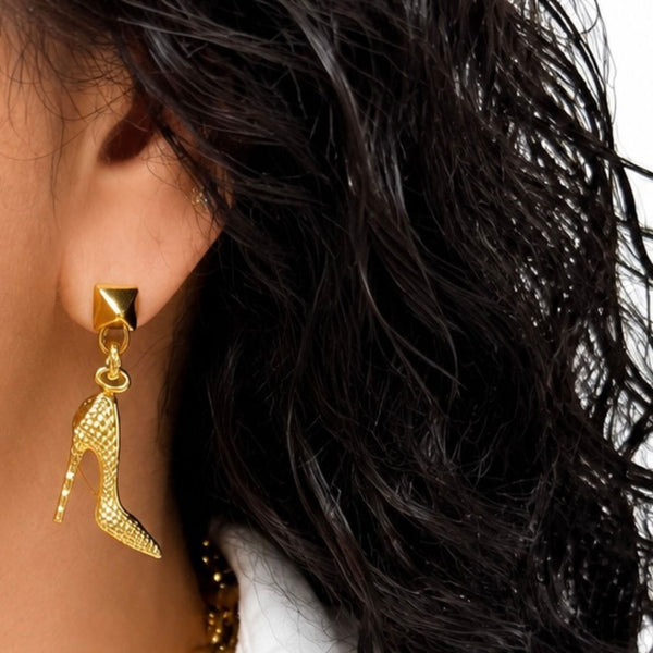 A close-up of a model's ear wearing a gold stud earring with a dangling high-heeled shoe charm, set against dark hair and a white collar.