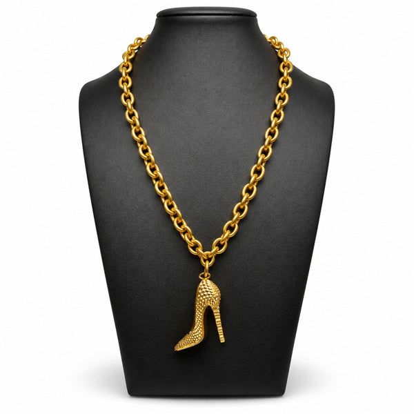 A gold chain necklace with a high-heeled shoe pendant, displayed on a black jewelry bust against a white background.