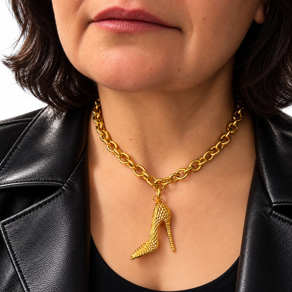 A close-up of a model wearing a textured gold high-heeled shoe pendant necklace on a gold link chain, styled with a black leather jacket.