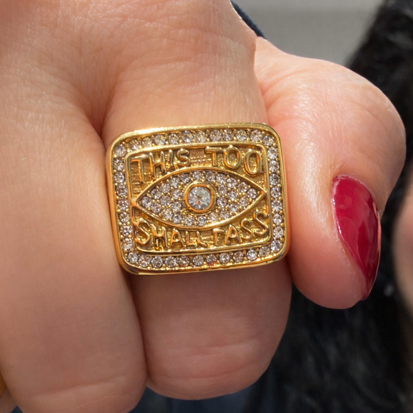 Gold ring with eye design and text on a finger with red nail polish.