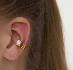 Gold earring with a clear gemstone worn by a person with brown hair.
