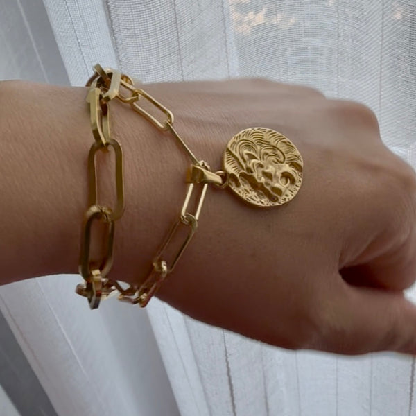 Gold chain bracelet with a round medallion on a wrist against a white curtain background