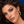 A close-up portrait of a woman with dark wavy hair wearing gold jewelry, including a gold hoop earring, a gold snake-shaped ear cuff, and a gold ring, against a blue background.