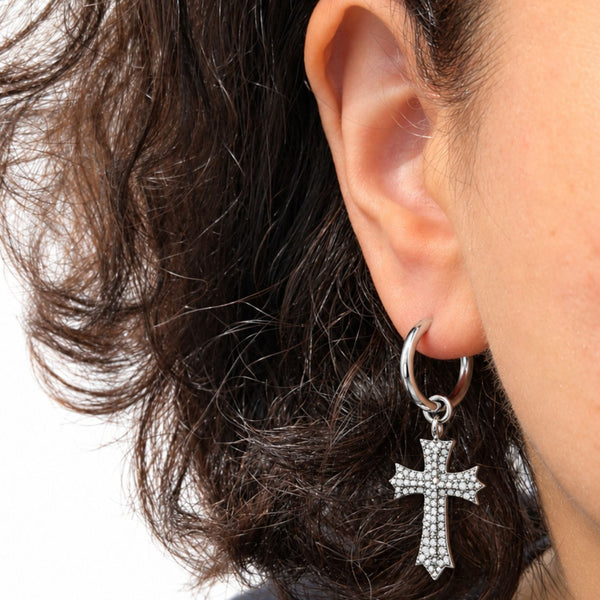 A close-up of a person's ear wearing a silver hoop earring with a dangling cross charm encrusted with small clear stones, against a white background.