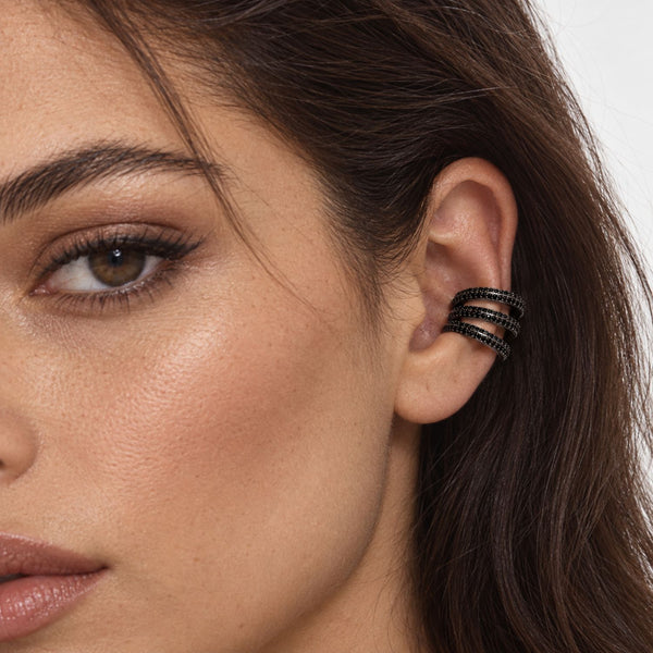 A close-up of a woman's ear wearing a black triple-band ear cuff with small clear stones, set against a clean white background.