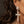 A close-up of a woman's ear and side profile wearing a large silver-toned drop earring with a pyramid stud and a cross-shaped pendant covered in clear stones.