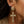A close-up of a model's ear wearing a gold cross drop earring adorned with clear crystals, alongside a small black stud and a round crystal earring.