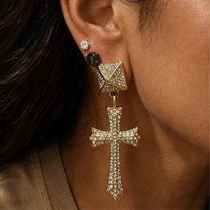 A close-up of a model's ear wearing a gold cross drop earring adorned with clear crystals, alongside a small black stud and a round crystal earring.