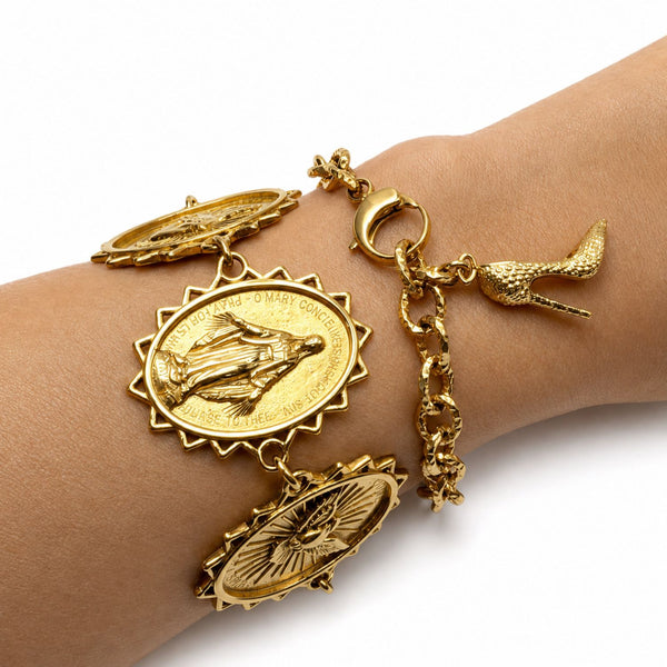 A gold charm bracelet with three religious medallion charms, a high-heeled shoe charm, and a sunburst bird charm, shown worn on a person's wrist against a white background.