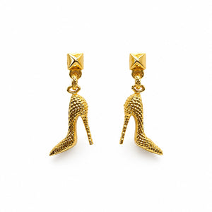 A pair of gold high-heeled shoe dangle earrings with textured details and pyramid stud tops, shown on a plain white background.
