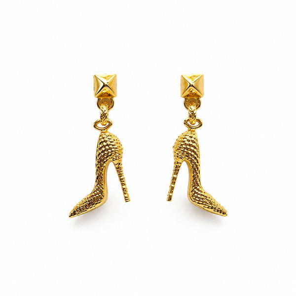 A pair of gold high-heeled shoe dangle earrings with textured details and pyramid stud tops, shown on a plain white background.