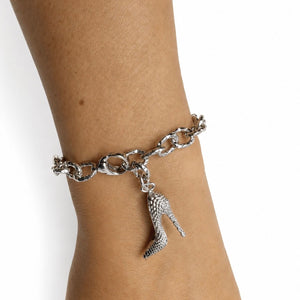 A silver-tone chain bracelet worn on a person's wrist, featuring a dangling high-heeled shoe charm with a textured, rhinestone-like surface.
