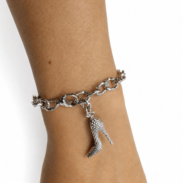A silver-tone chain bracelet worn on a person's wrist, featuring a dangling high-heeled shoe charm with a textured, rhinestone-like surface.