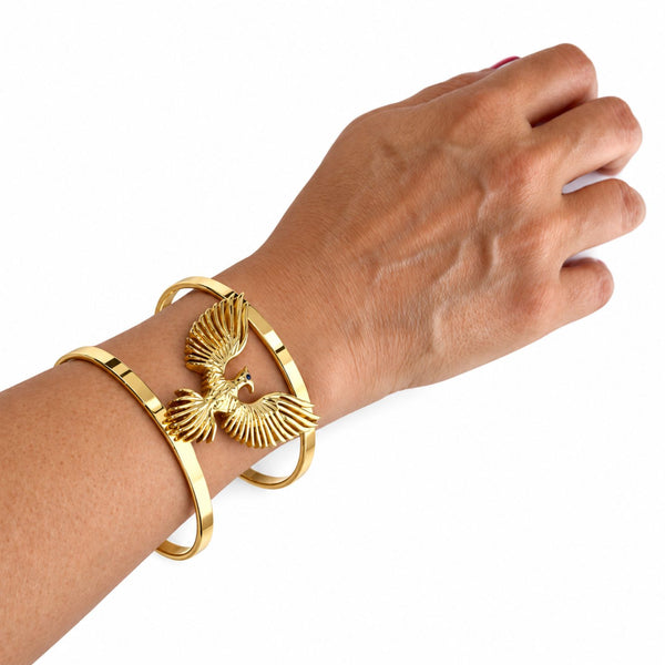 A gold-toned cuff bracelet with a raised eagle motif worn on a person's wrist, shown against a plain white background.