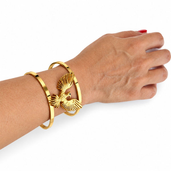 A close-up of a person's wrist wearing two gold-toned bracelets, including a bangle with a raised bird motif, against a plain white background.