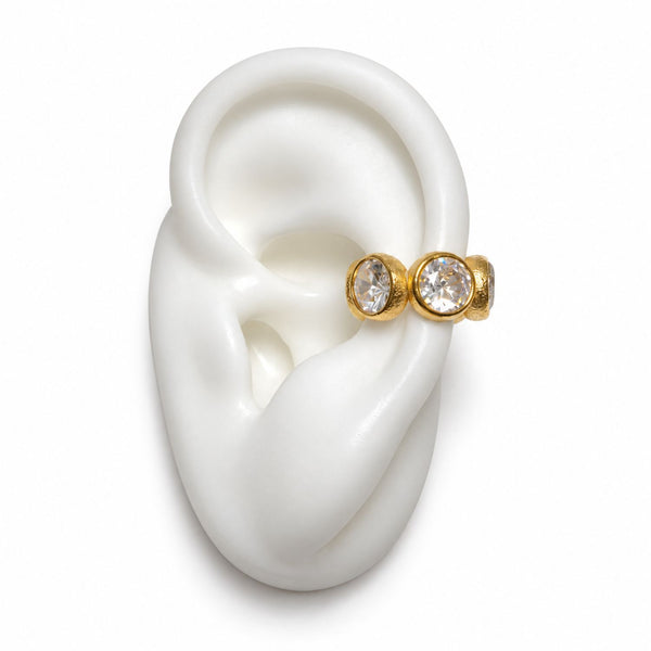 A pair of gold ear cuff rings with clear round gemstones, displayed on a white ear mannequin against a white background.