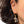 A close-up of a person's ear wearing a gold huggie hoop earring with a dangling cross charm paved with small clear stones.