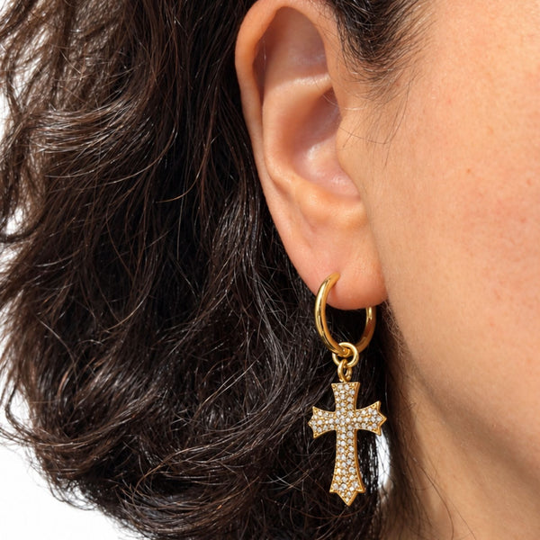 A close-up of a person's ear wearing a gold huggie hoop earring with a dangling cross charm paved with small clear stones.