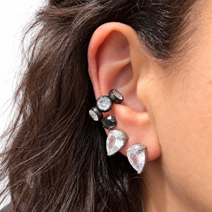 A close-up of a person's ear wearing a cluster of black ear cuff studs with clear faceted gemstones, including pear-shaped and round-cut stones.