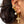 A close-up of a person's ear wearing a stack of gold ear piercings, including crystal-studded hoops, a skull stud, a pearl drop, and a dangling cross charm.