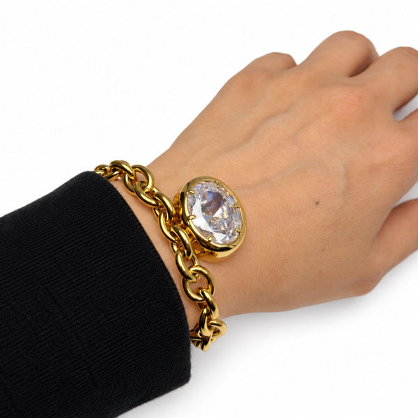 A gold chain bracelet with a large oval clear crystal pendant, shown worn on a person's wrist with a black sleeve visible.